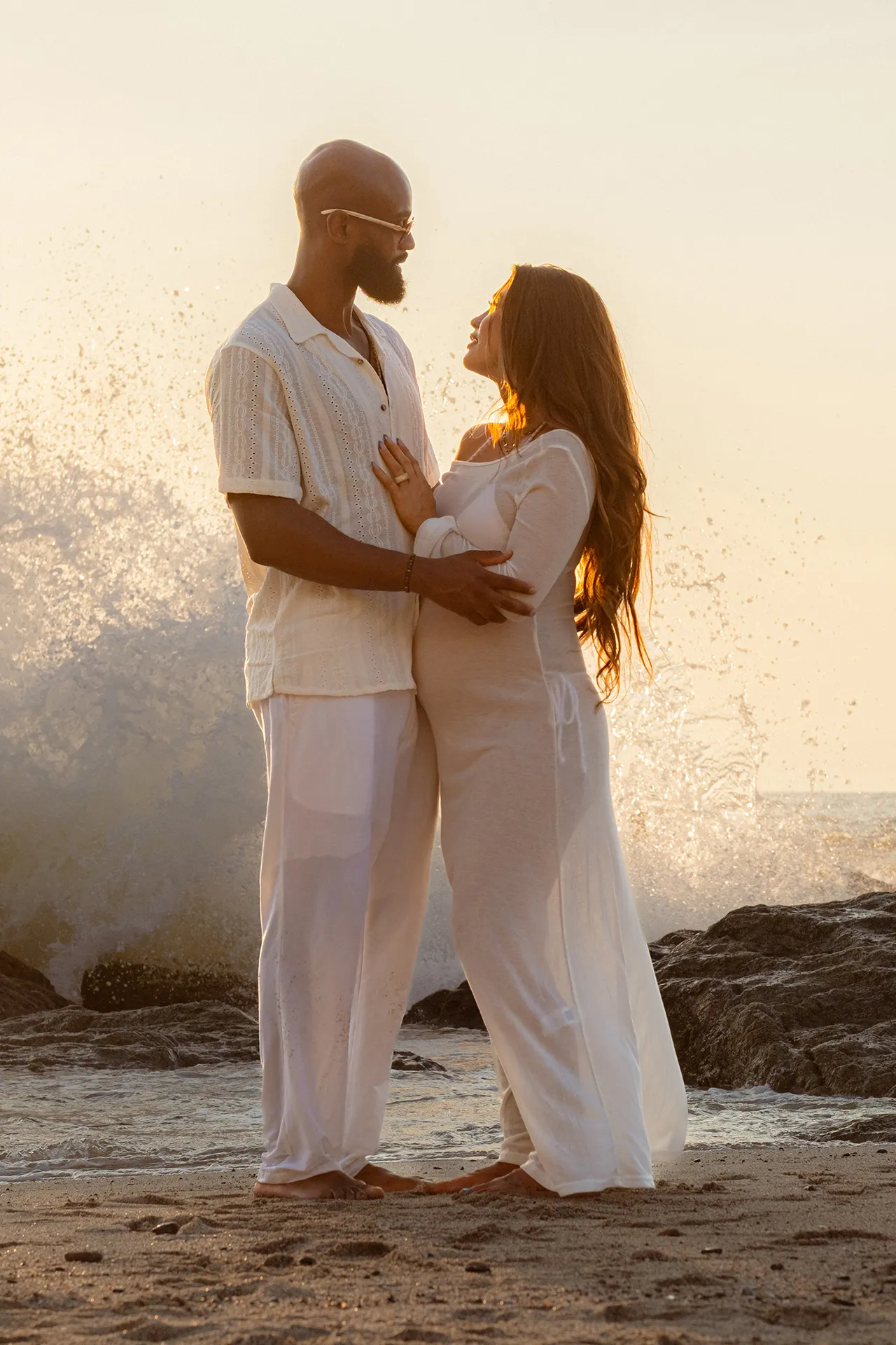 Couple dressed in white facing each other at sunset, his hand resting on the baby bump as a wave breaks behind them — babymoon maternity session in Puerto Vallarta